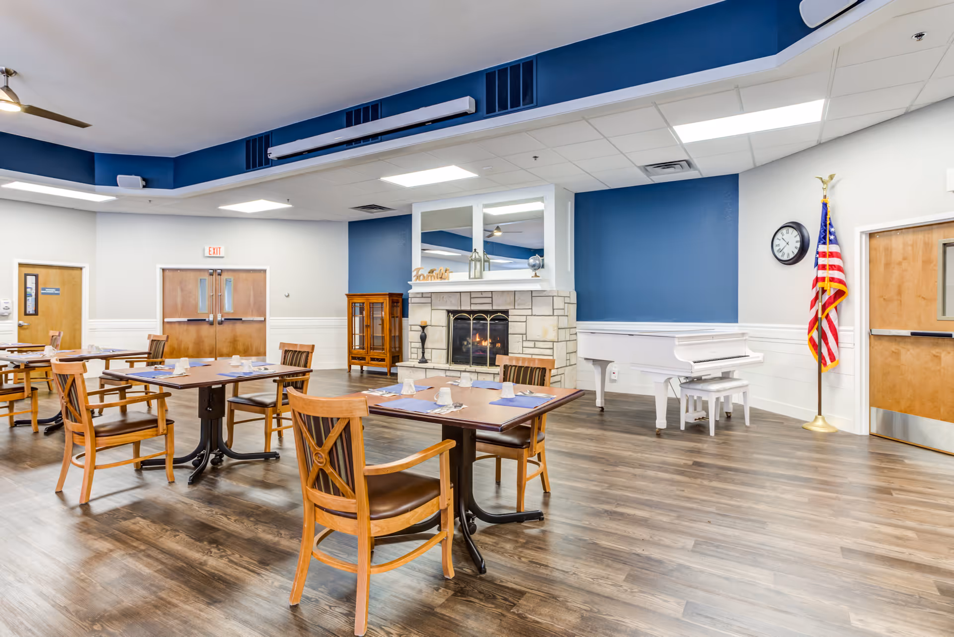 A spacious dining room with wooden tables and chairs set with placemats, cups, and utensils. The room features a stone fireplace with a mirror above it, a white grand piano, an American flag, and a clock on the wall. The walls are painted blue and white, and the floor is wood.