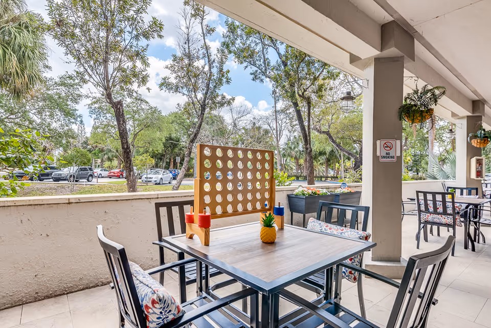 Outdoor covered patio area at a senior living facility with tables and chairs. One table has a large wooden Connect Four game and a small pineapple decoration. There are hanging plants and a 'No Smoking' sign on a pillar. Trees and parked cars are visible beyond the patio.