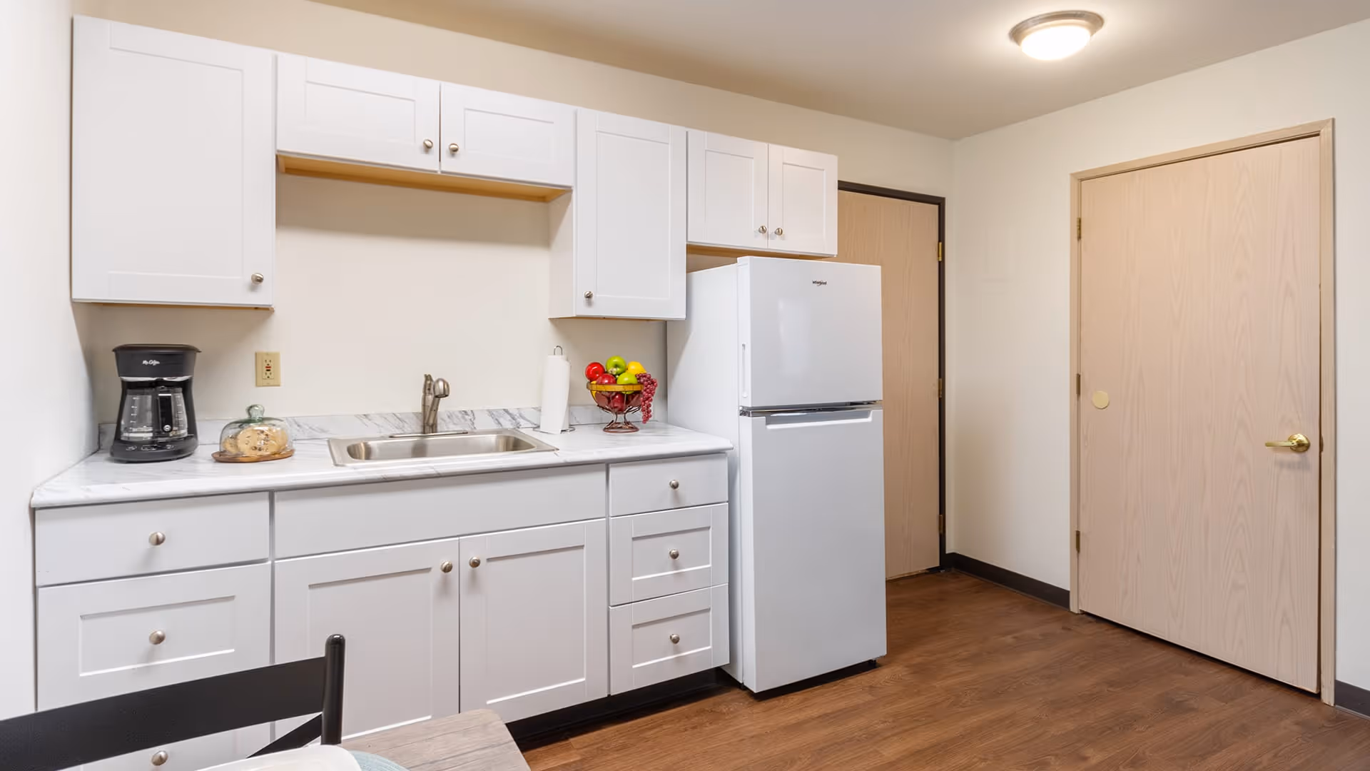 A clean and simple kitchen area with white cabinets, a white refrigerator, a stainless steel sink, a coffee maker, a paper towel holder, and a bowl of fruit on the countertop. The floor is wood, and there are two closed wooden doors in the background.