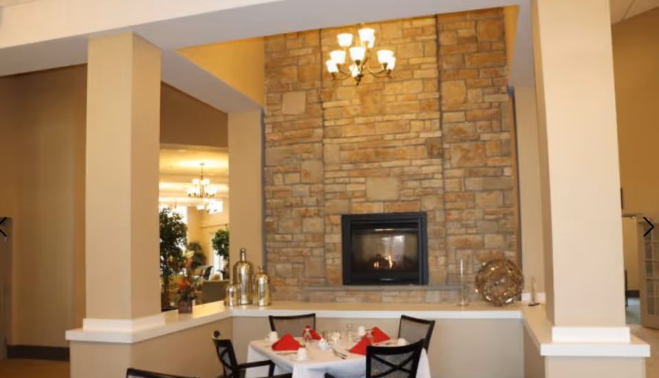 A cozy dining area with a table set for four, featuring white tablecloth, red napkins, and dining ware. The table is positioned in front of a large stone fireplace with a lit fire. The room has beige walls and pillars, with decorative items on the ledge around the fireplace and a chandelier hanging from the ceiling.