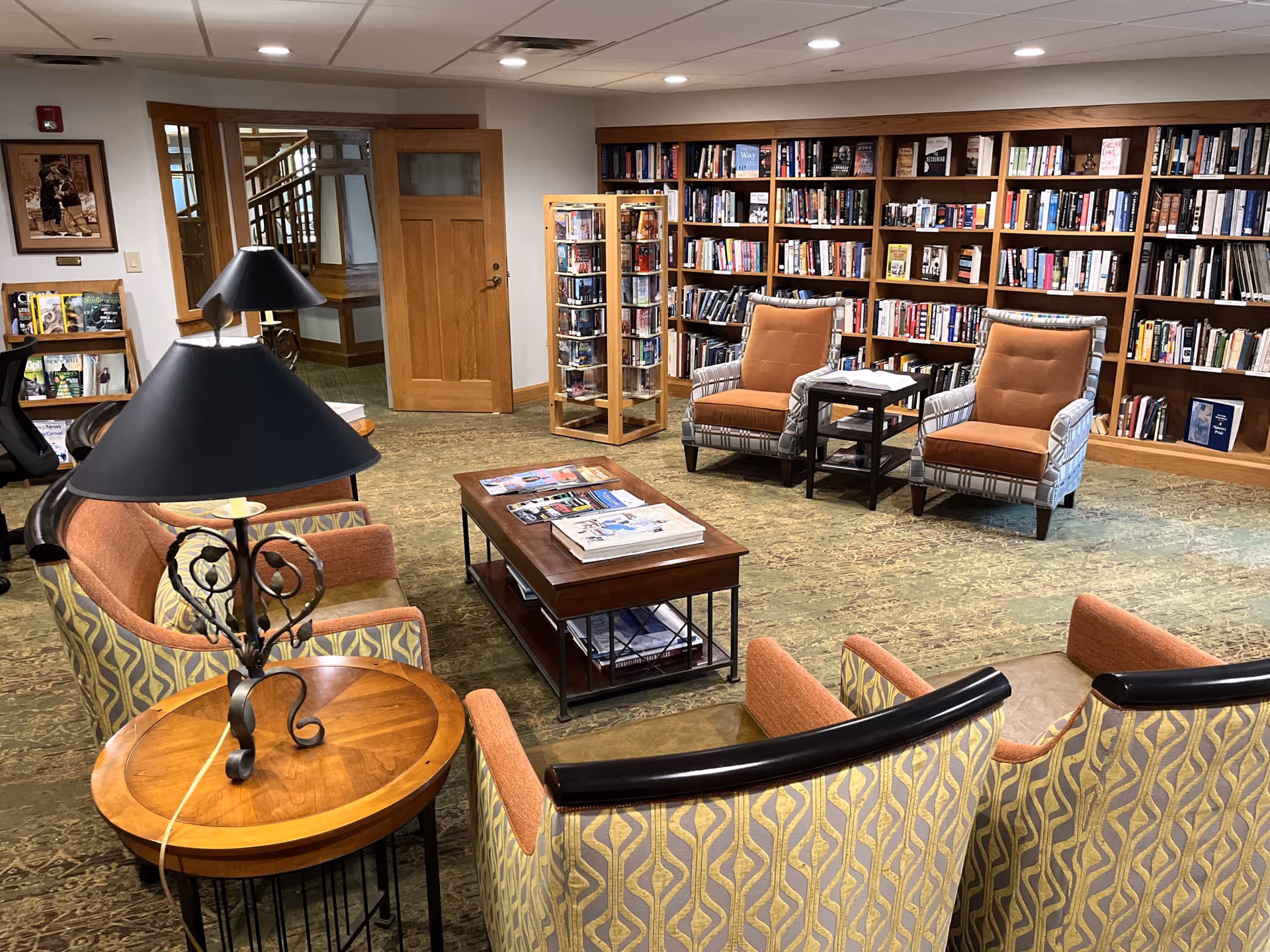 A cozy library or reading room with bookshelves filled with books along the back wall, two cushioned armchairs with orange seats and patterned backs, a wooden coffee table with magazines and books, and several other cushioned chairs arranged around the table. There are also two black lamps on wooden side tables, a wooden door, and a carpeted floor.