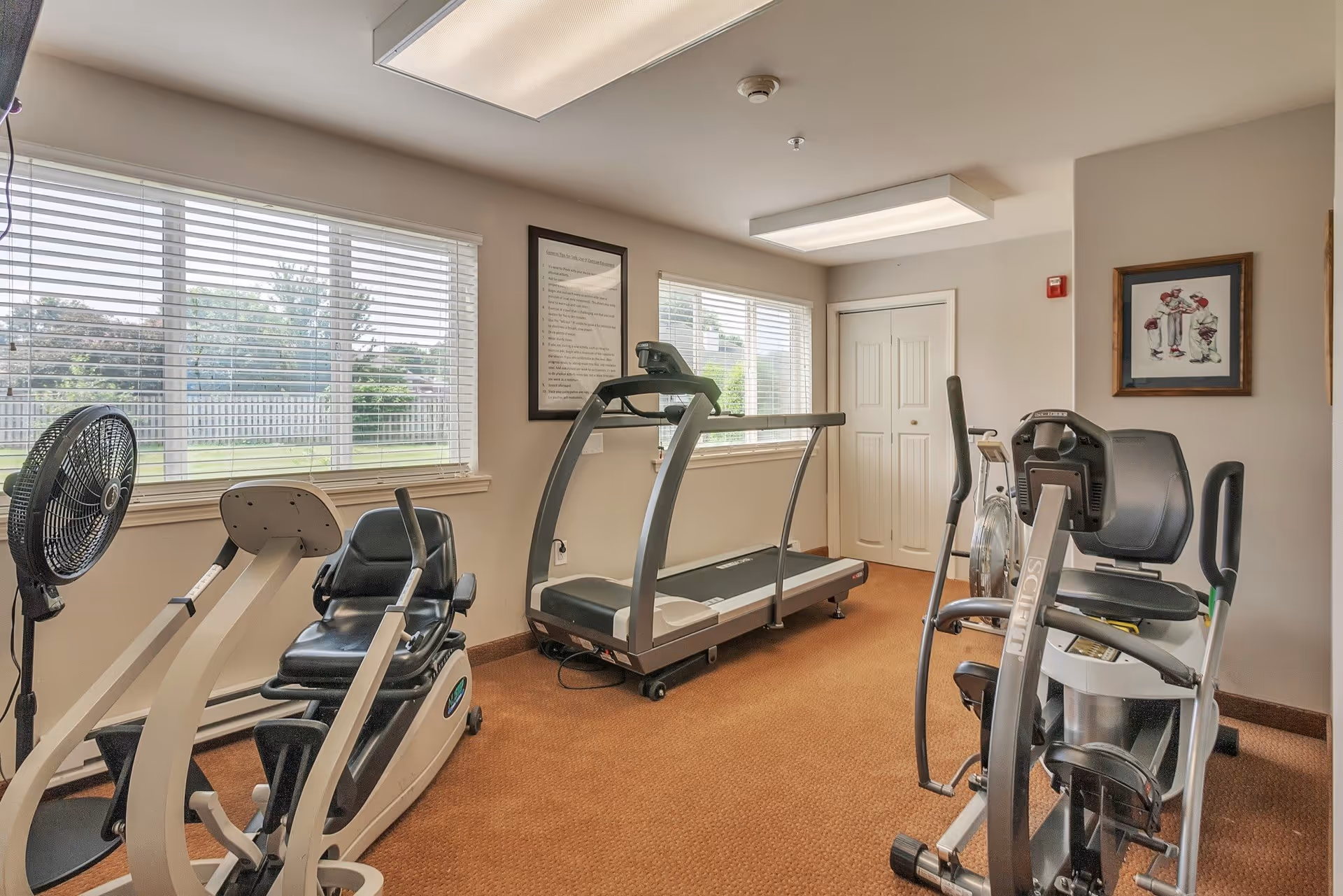 Small exercise room with a treadmill, stationary bikes, and a fan. The room has beige walls, two large windows with white blinds letting in natural light, framed pictures on the walls, and a brown carpeted floor.