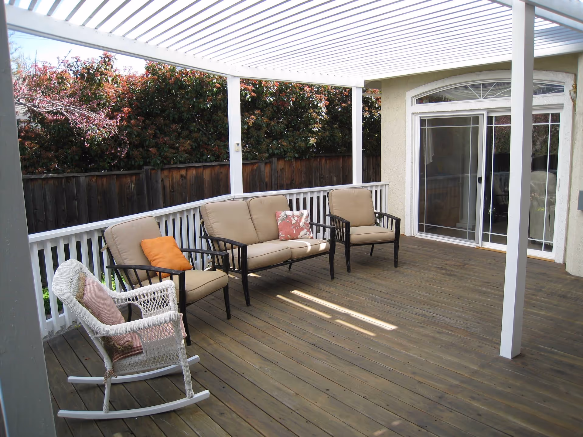 Covered wooden patio with outdoor seating including a white rocking chair with a cushion, two cushioned armchairs, and a cushioned loveseat with pillows. The patio has a white railing and a wooden fence with greenery behind it. Sliding glass doors lead inside the building.