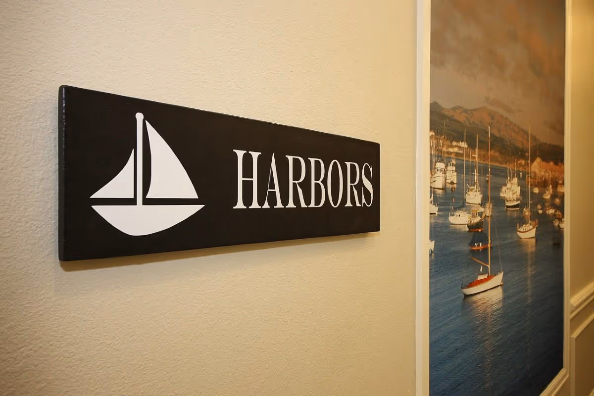 A black rectangular sign mounted on a beige wall with a white sailboat icon and the word 'HARBORS' in white capital letters. To the right, there is a large framed picture showing a harbor scene with multiple sailboats on the water and mountains in the background.