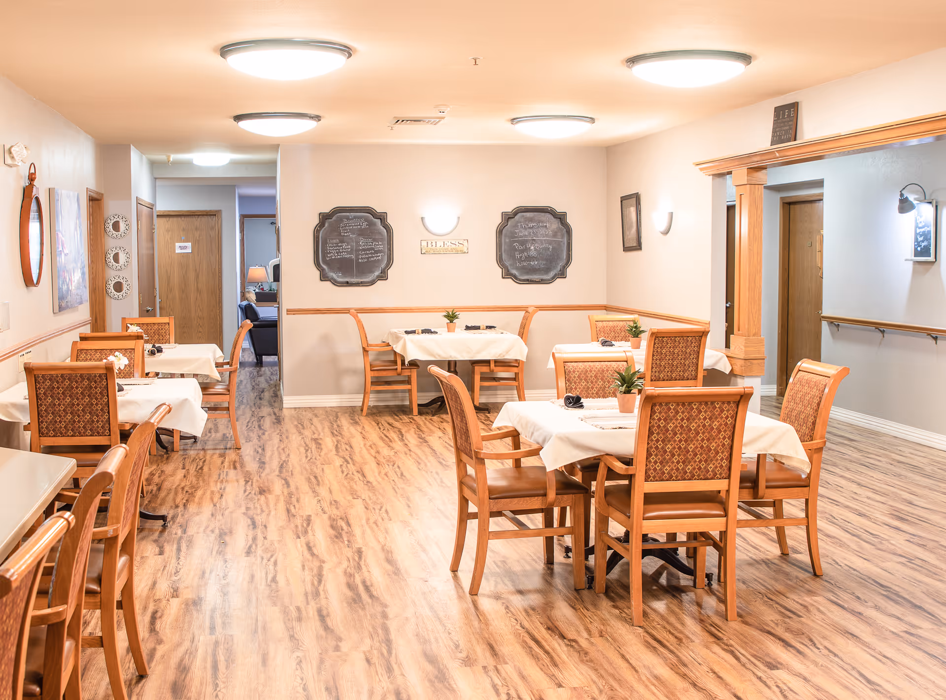 Bright dining room with several set tables and wooden chairs on wood-look flooring in an assisted living facility.
