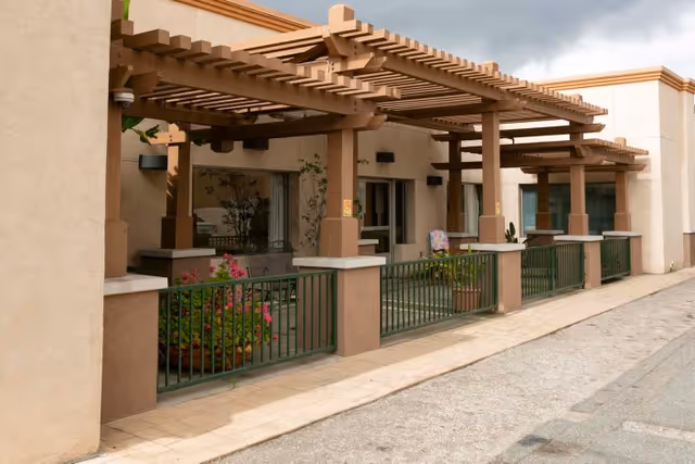 Outdoor patio area with wooden pergolas attached to a beige building. The patio is enclosed by green metal railings and has potted plants and flowers. The sky is cloudy.