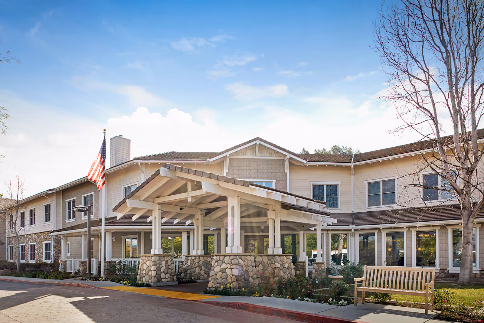 Exterior front view of a senior living facility building with a covered entrance supported by stone pillars, an American flag on a flagpole, a wooden bench, and leafless trees under a partly cloudy sky.