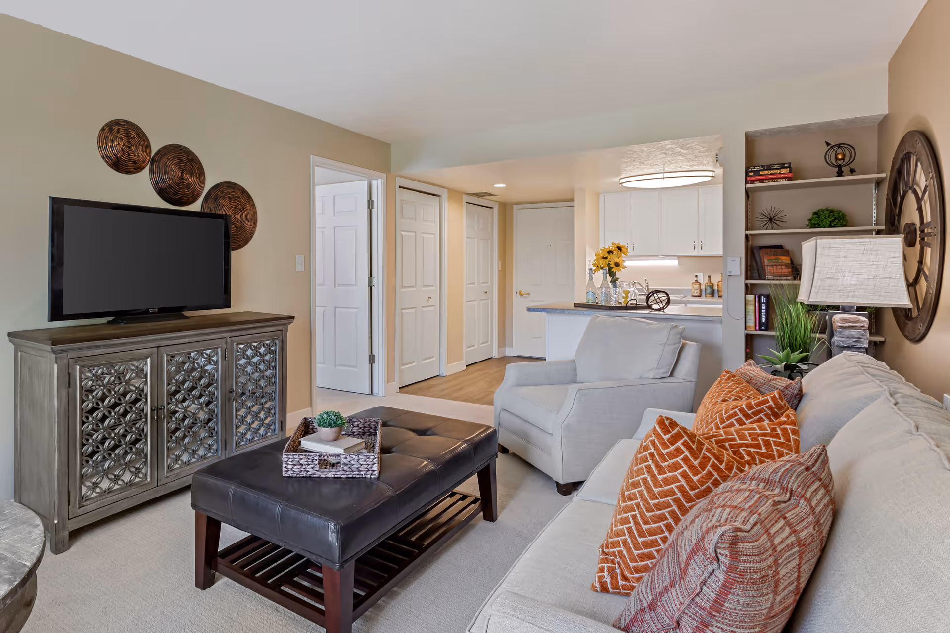A cozy living room with a beige sofa adorned with orange and patterned pillows, a gray armchair, and a dark leather ottoman serving as a coffee table with a tray and small plant on it. A TV sits on a decorative cabinet against the wall, with three round wall decorations above it. In the background, there is a kitchen area with white cabinets, a countertop with flowers and decorative items, and a bookshelf with books and decor. The room has beige walls and carpeted flooring.