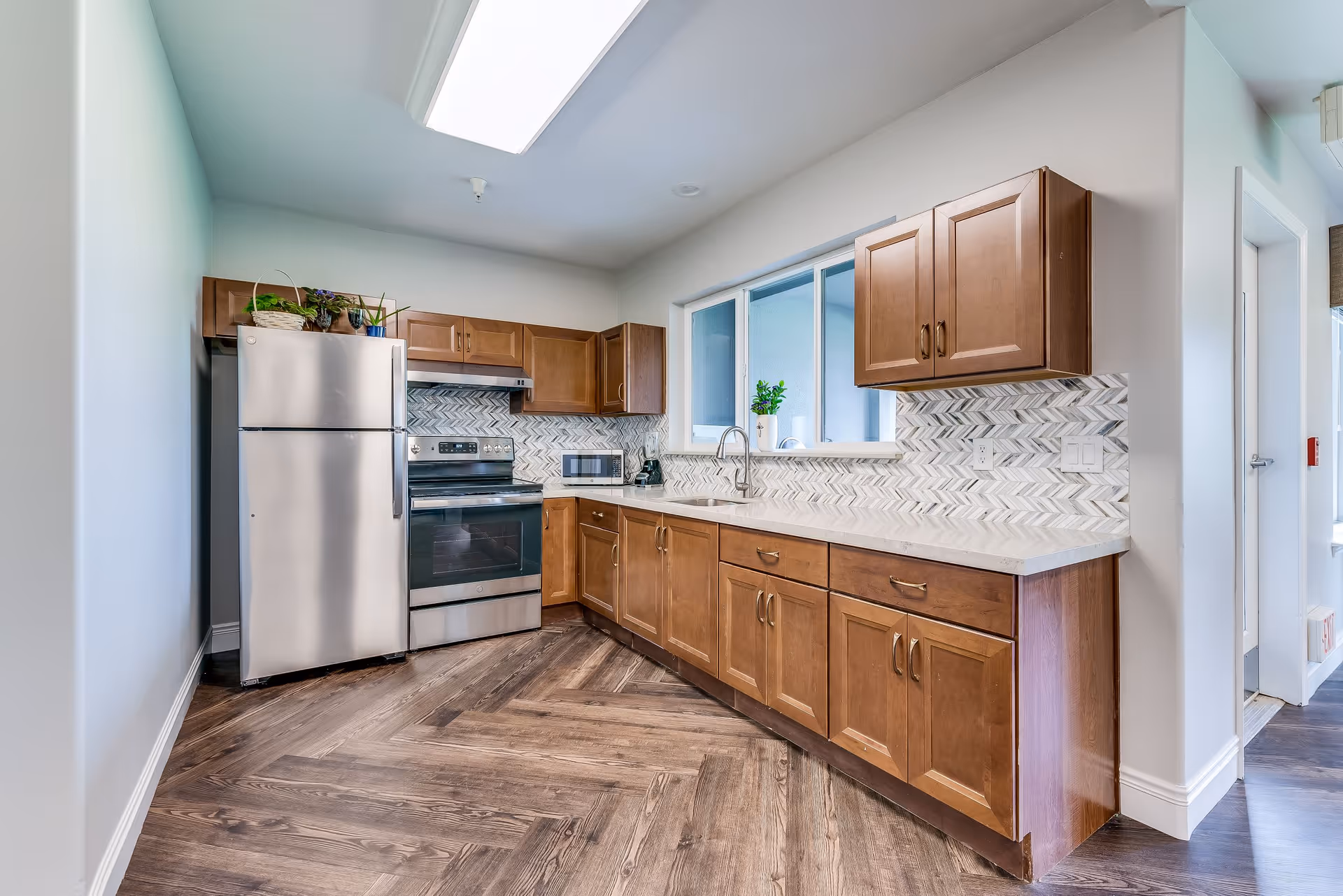 A modern kitchen with wooden cabinets, a stainless steel refrigerator and oven, a microwave, and a sink under a window. The backsplash features a chevron tile pattern, and the floor has a wood-like finish arranged in a herringbone pattern.