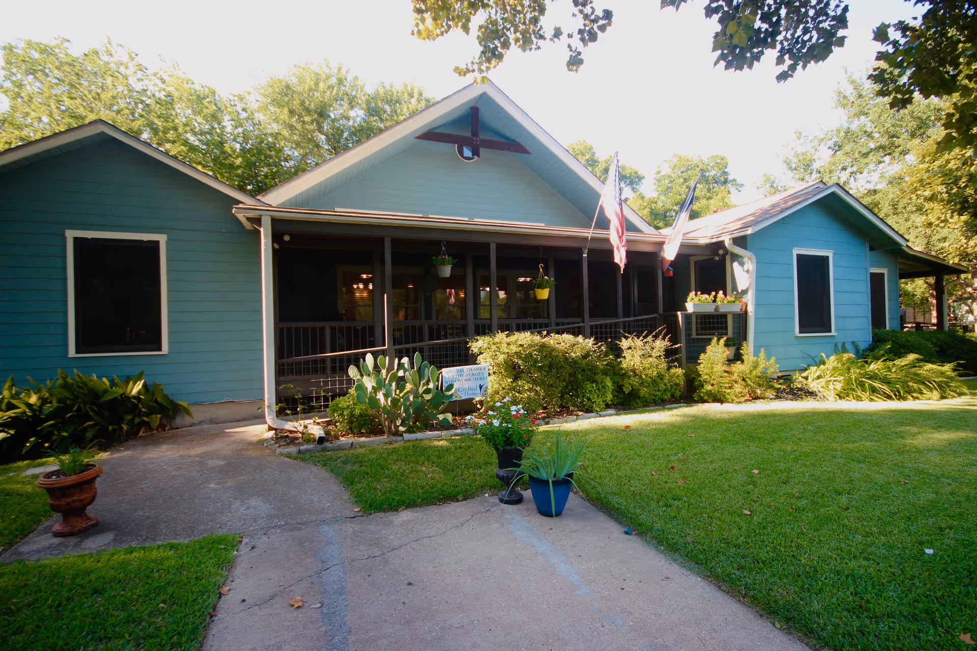 Exterior view of a single-story blue house with a screened porch, two American flags, and a well-maintained lawn with various plants and shrubs in front.