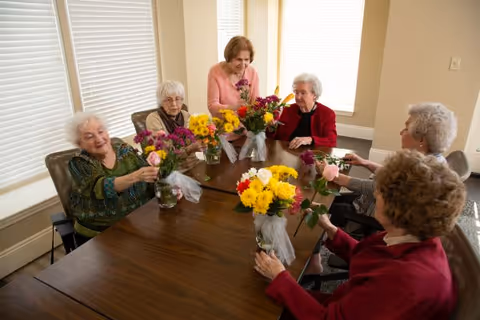 Six elderly women seated around a wooden table arranging colorful flower bouquets in a bright activity/dining room.