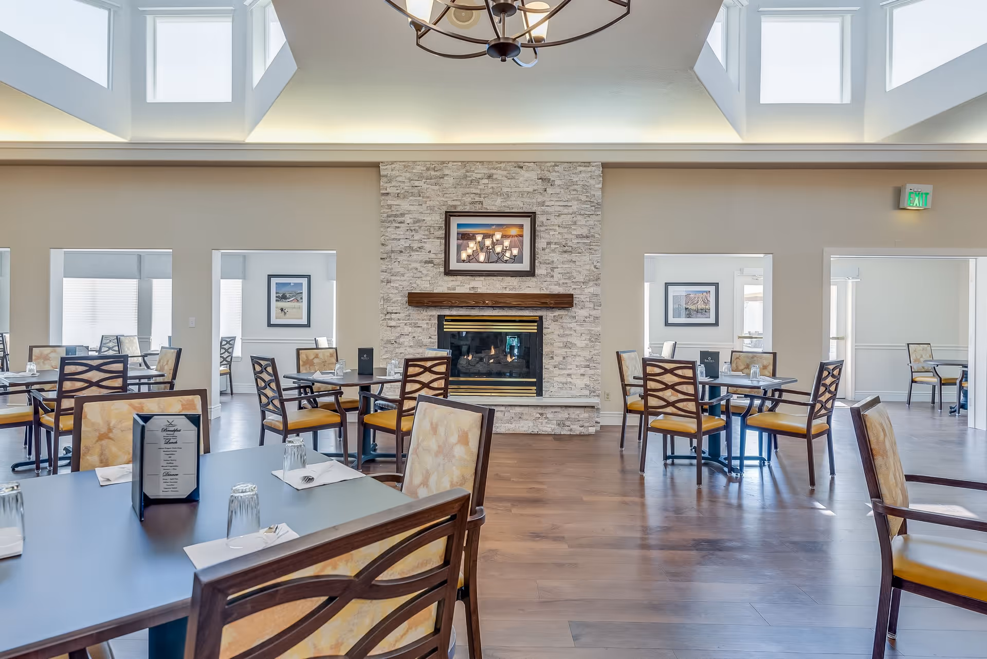A bright dining room with multiple tables and chairs arranged neatly. The room features a stone fireplace with a framed picture above it, large windows near the ceiling allowing natural light to fill the space, and hardwood flooring. Each table is set with napkins, utensils, and upside-down drinking glasses.