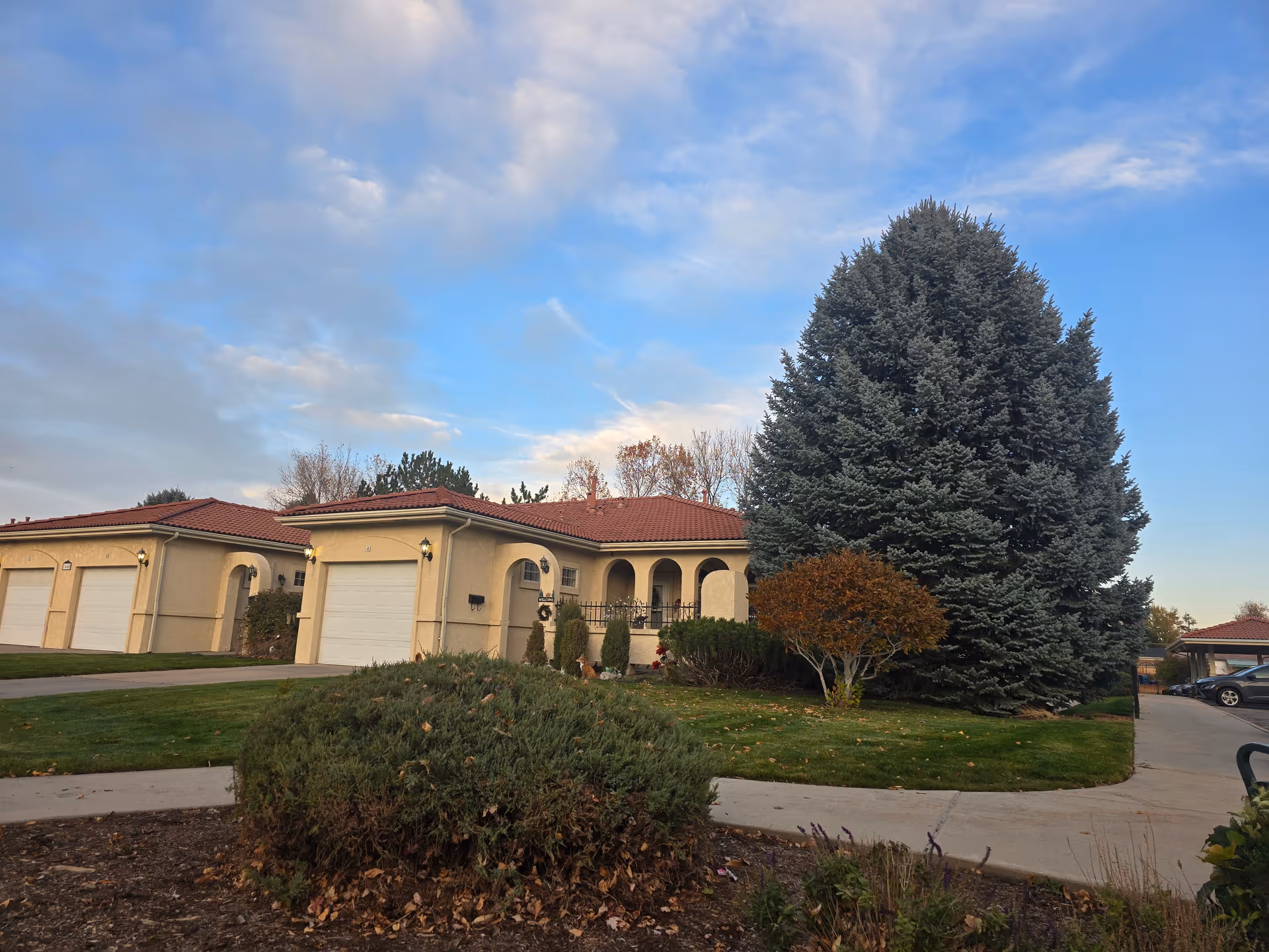 Exterior view of a single-story residential building with beige walls and a red-tiled roof, surrounded by green grass, bushes, and a large evergreen tree under a partly cloudy blue sky.