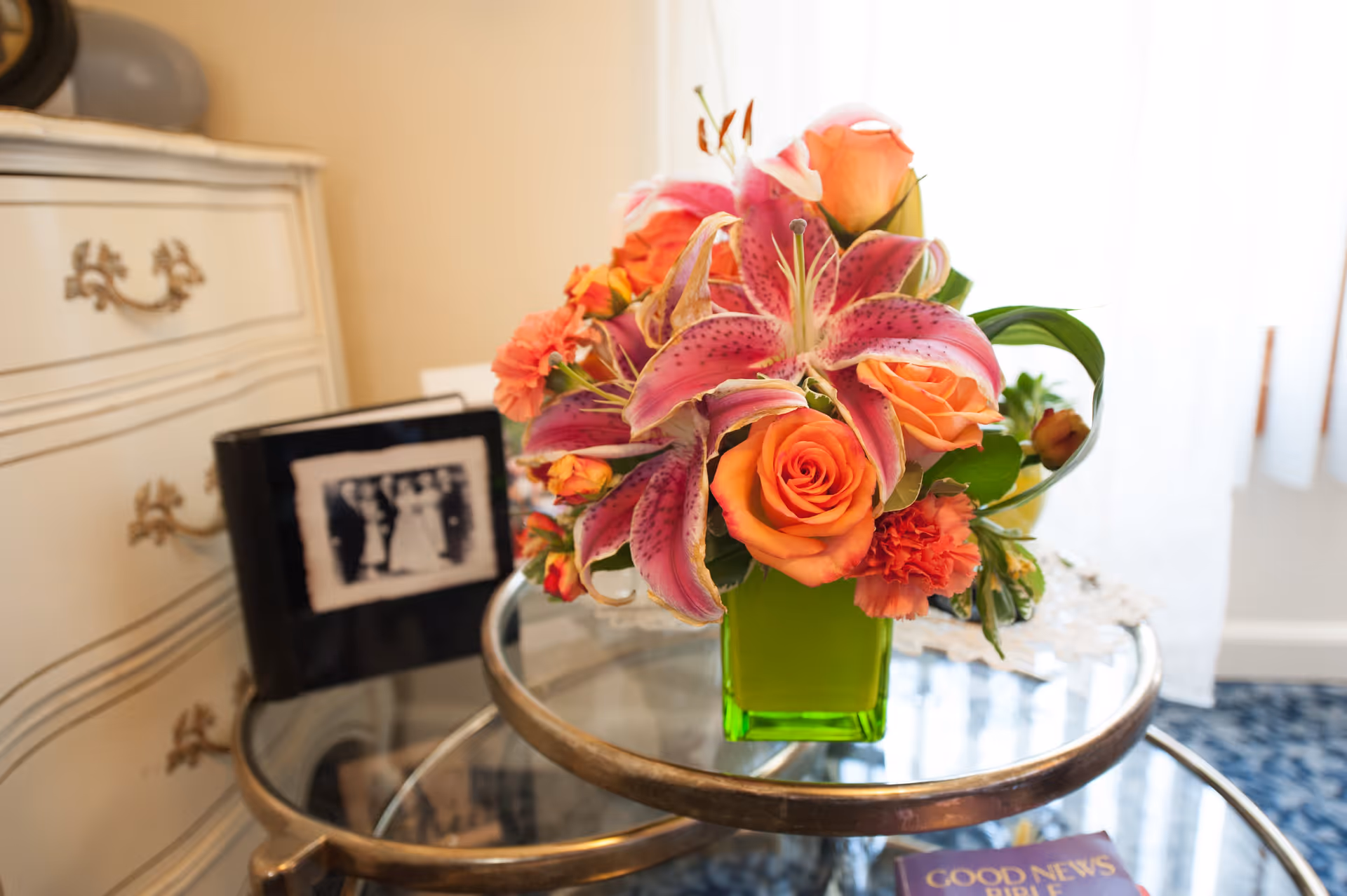 A close-up of a vibrant floral arrangement with pink lilies and orange roses in a green vase on a glass-topped round table. In the background, there is a white dresser with ornate handles and a framed black and white photo. A book titled 'Good News Bible' is partially visible on the table.