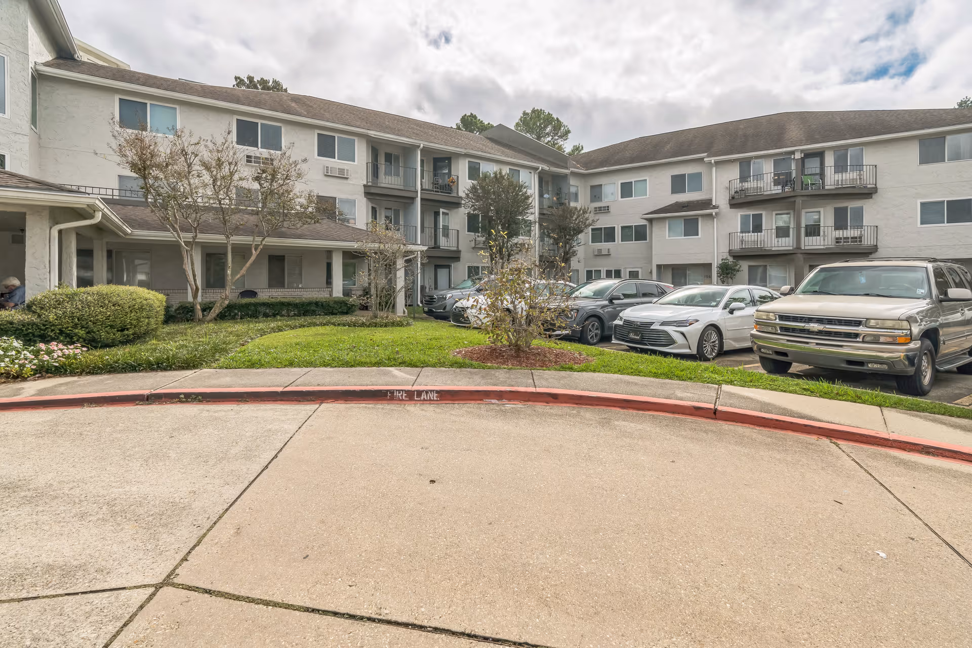 Front exterior of a three-story senior living building with balconies, landscaped lawn, parked cars, and a circular driveway.