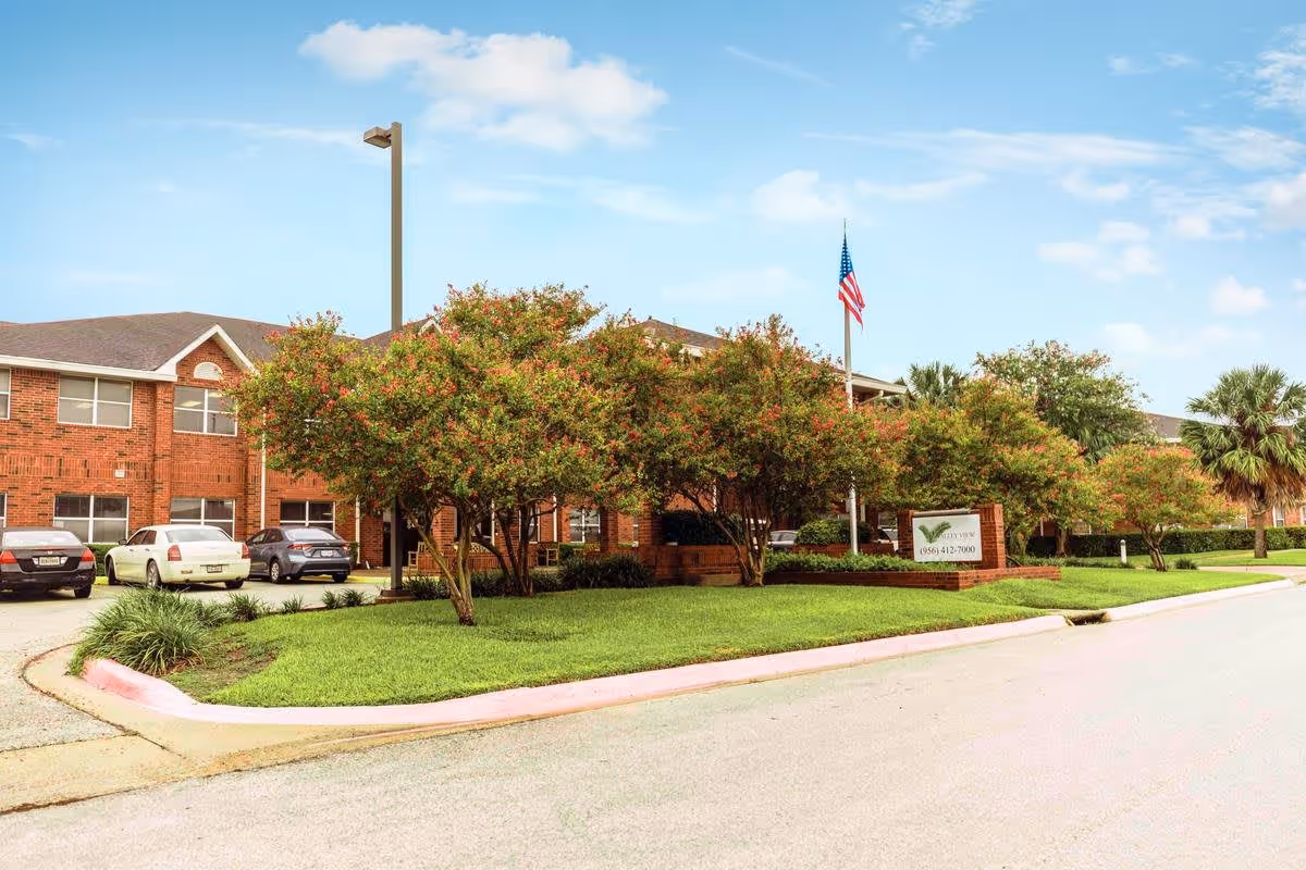 Front view of a two-story brick senior living building with landscaped lawns, trees, parked cars, and an American flag.