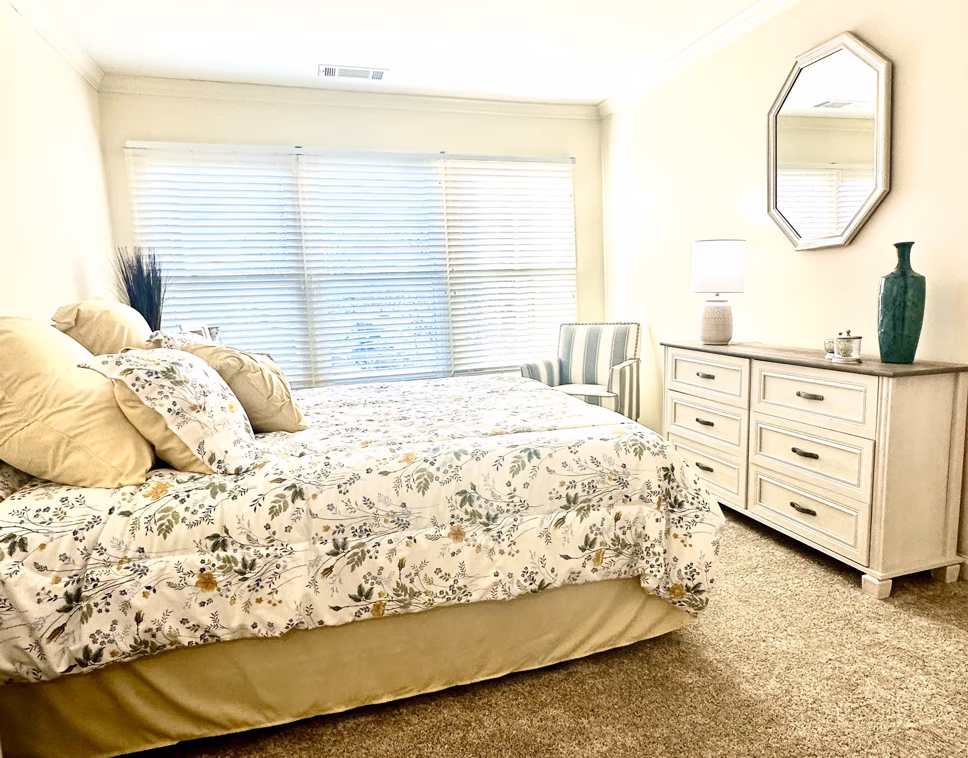 Bright bedroom with a floral-covered bed, dresser with mirror and lamp, and a striped armchair by a large window.