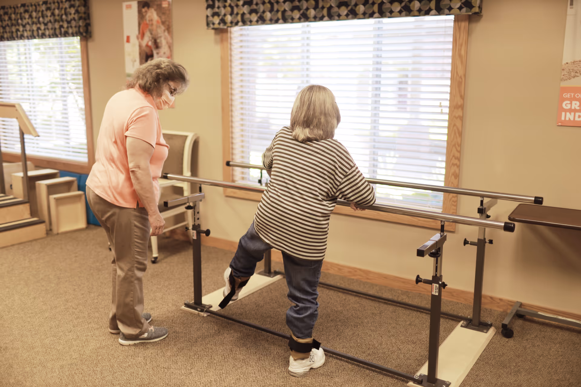 An elderly woman wearing a striped shirt and jeans is performing a leg exercise using parallel bars in a rehabilitation or physical therapy room. Another elderly woman, wearing a peach shirt and beige pants with a face mask, is standing nearby observing or assisting. The room has large windows with blinds and patterned valances, and exercise equipment is visible in the background.