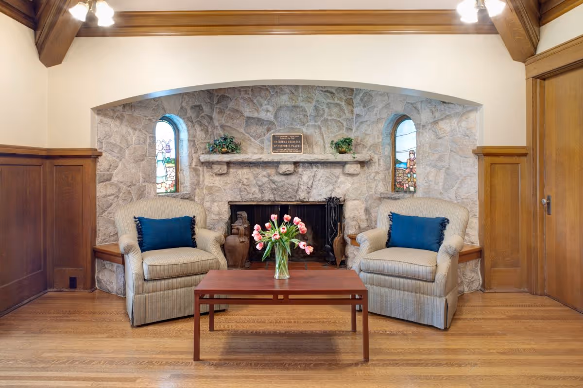 A cozy living room area featuring two beige armchairs with blue cushions placed on either side of a wooden coffee table with a vase of pink tulips. Behind the chairs is a stone fireplace with a wooden mantel, flanked by two small stained glass windows. The room has wooden paneling on the walls and hardwood flooring.
