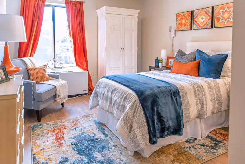 A bright and cozy bedroom featuring a neatly made bed with white and gray striped bedding, blue and orange accent pillows, and a blue throw blanket. The room includes a colorful area rug, a gray armchair with an orange pillow and white throw blanket, a white wardrobe, a wooden dresser with a lamp, and three vibrant framed artworks on the wall above the bed. A window with red curtains allows natural light to fill the space.
