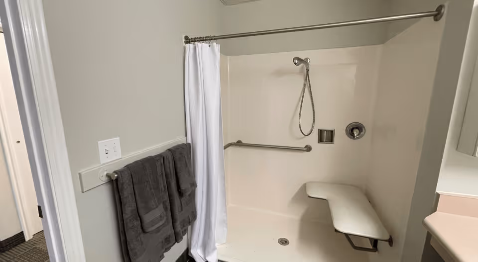 A bathroom shower area with a white shower curtain, a built-in foldable bench, a handheld showerhead, and grab bars. Two dark gray towels hang on a towel rack outside the shower. The walls are light-colored, and part of a countertop with a mirror is visible on the right side.
