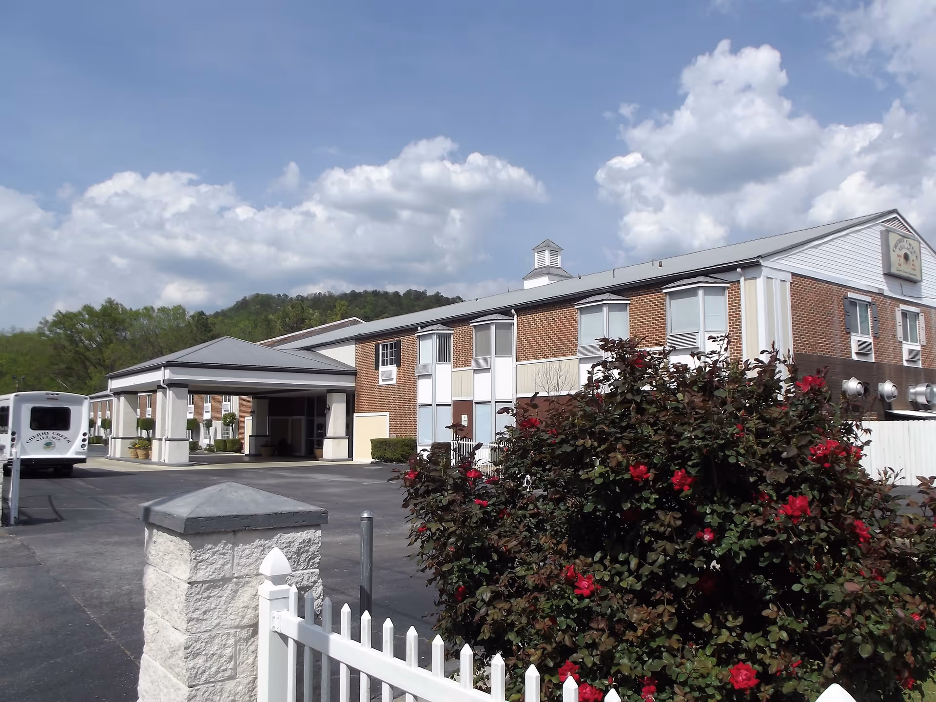 Brick senior living facility with entrance canopy, driveway, bus, and blooming roses in foreground.
