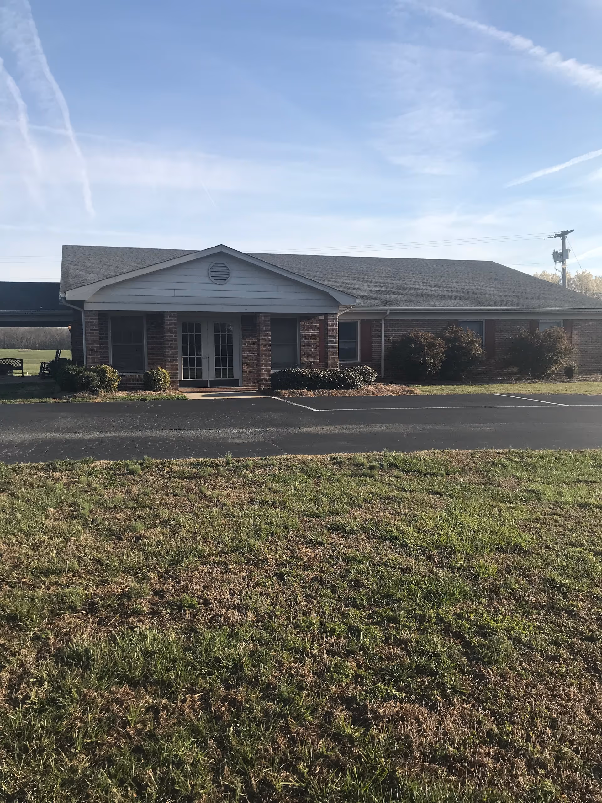Single-story brick building with a gray roof and white trim, featuring a covered entrance with double glass doors. The building is surrounded by a grassy lawn and a paved driveway or parking area in front. The sky is clear with some wispy clouds.