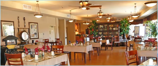 A spacious dining room with multiple tables covered in beige tablecloths, each set with cups, plates, and floral centerpieces. Wooden chairs surround the tables, and the room is warmly lit with ceiling lights and fans. Large windows on the right side allow natural light to fill the space, and there are plants and decorative items along the walls.