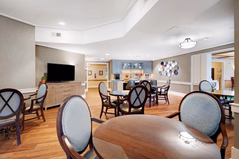 A bright and spacious common area in a senior living facility featuring multiple round wooden tables with cushioned chairs. A flat-screen TV is mounted on a wooden cabinet against the wall. The room has wood flooring, neutral-colored walls, and a decorative wall display of plates. In the background, there is a kitchenette area with blue cabinets and a countertop with flowers and snacks.