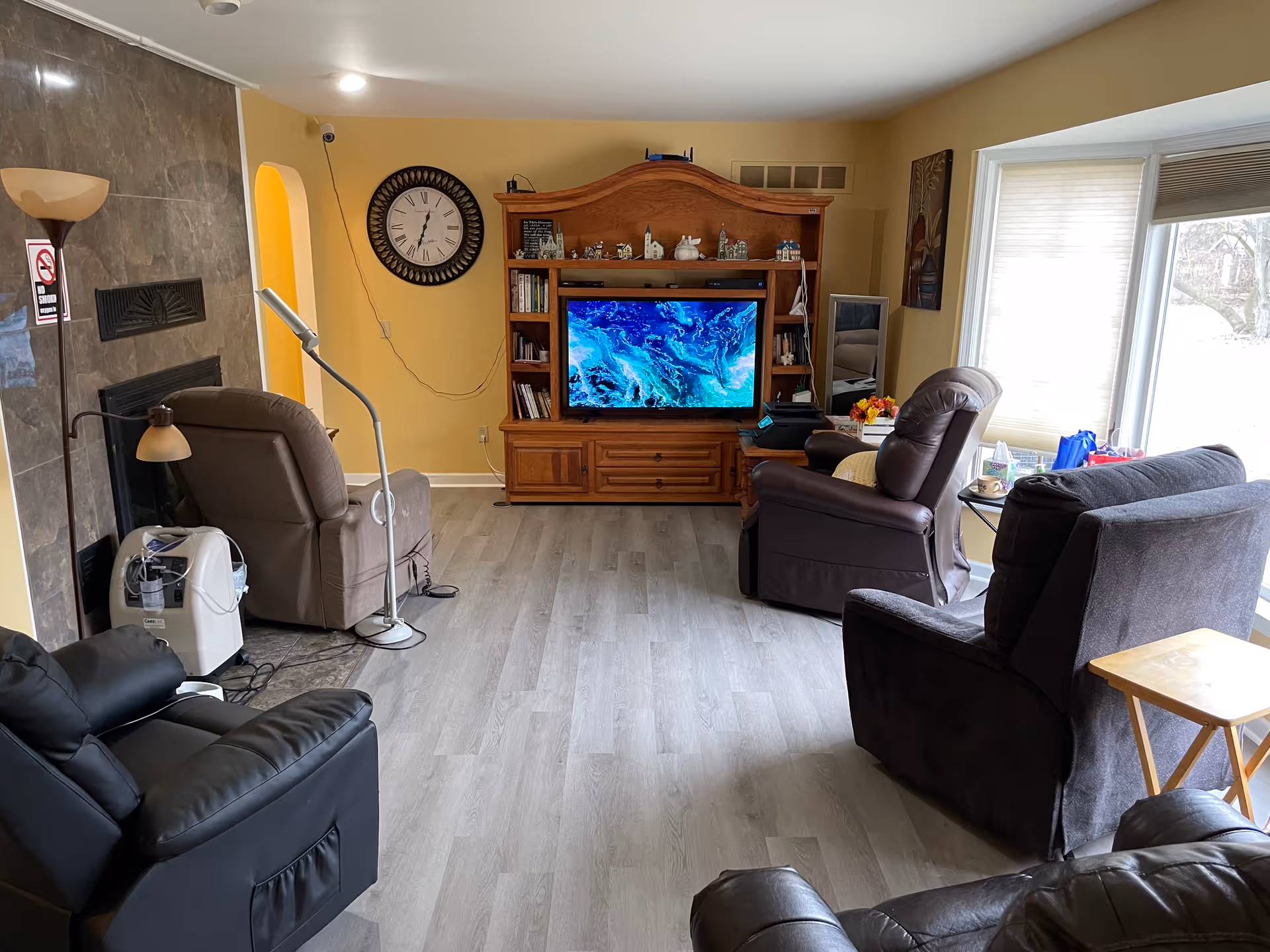 A cozy living room with four recliner chairs arranged around a wooden entertainment center with a large flat-screen TV displaying a blue abstract image. The room has light wood flooring, a fireplace with a no smoking sign, a standing lamp, a large wall clock, and a window with blinds letting in natural light. There are small decorative items on the entertainment center and a side table with tissues and other items near one of the chairs.