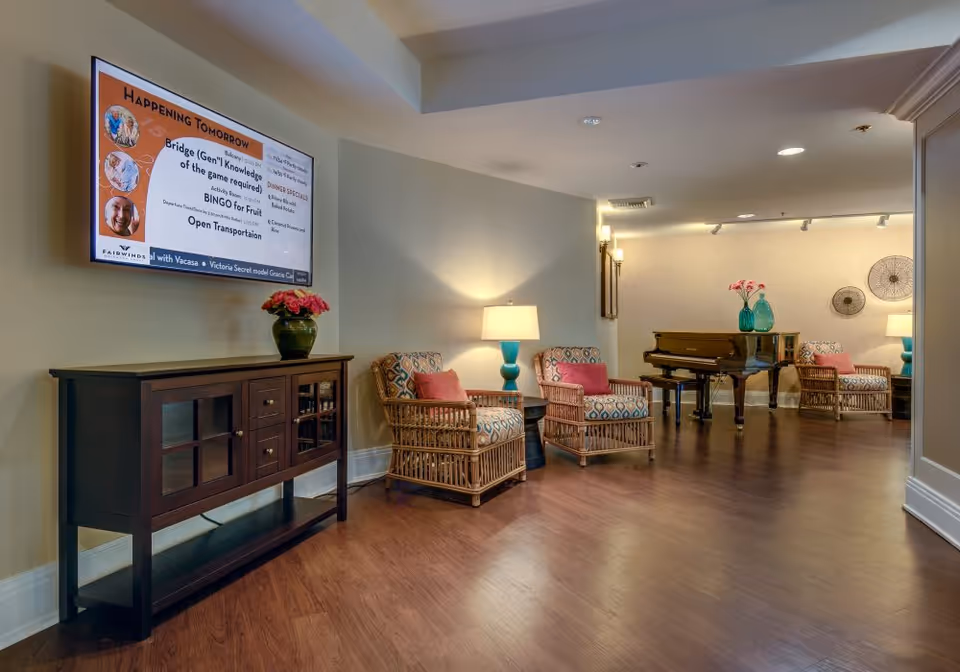 A cozy common area in a senior living facility featuring a wooden floor, a dark wooden console table with a green vase of pink flowers, two wicker armchairs with patterned cushions and pink pillows, a blue table lamp, and a grand piano in the background. A wall-mounted screen displays upcoming activities and dinner specials.