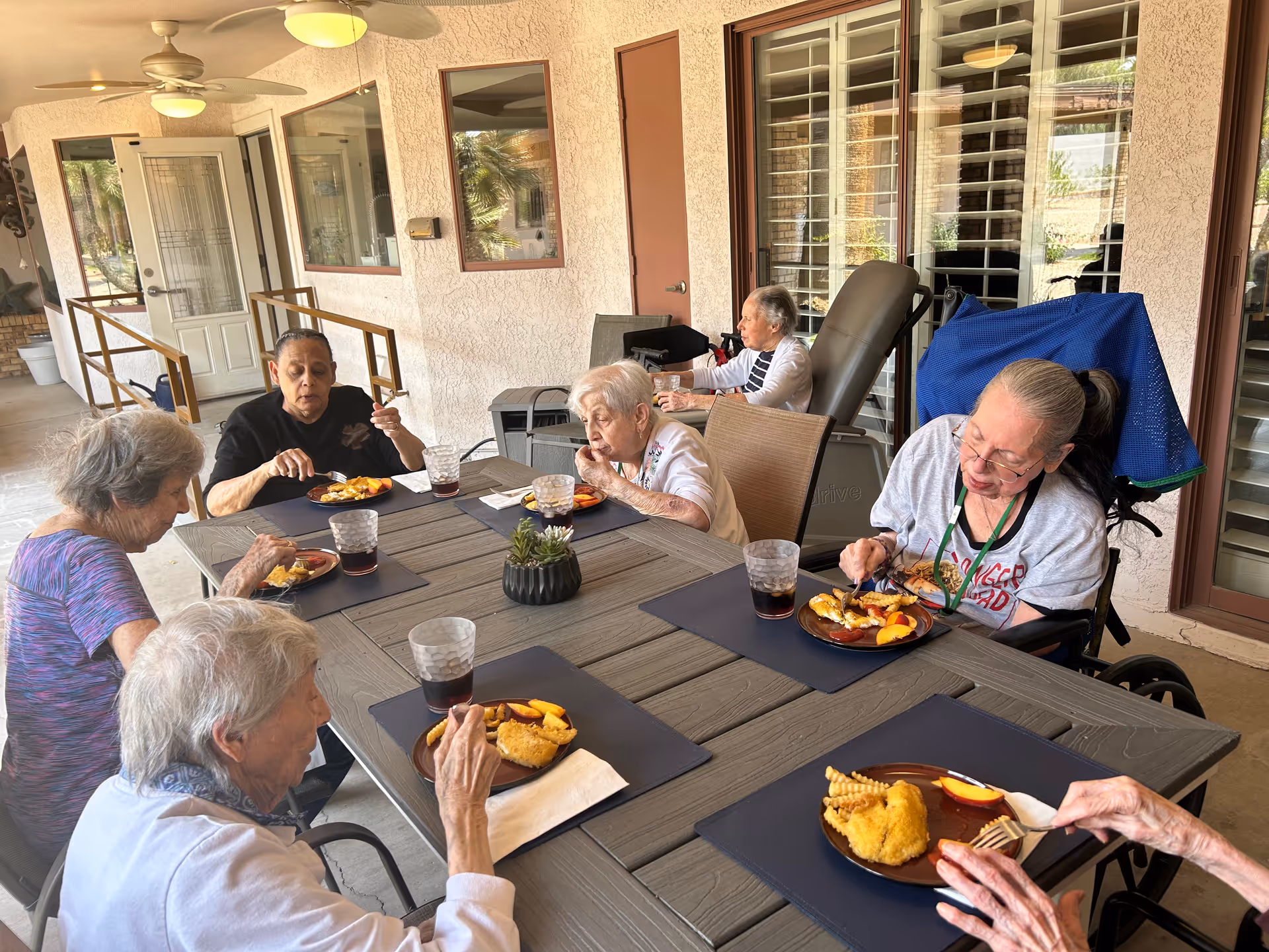 Six elderly residents sit around a covered outdoor patio table eating meals.