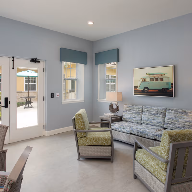 A cozy living room with light blue walls, two windows with blue valances, a framed picture of a vintage van with surfboards on top, a patterned blue sofa, two green cushioned armchairs, a side table with a lamp, and glass doors leading to an outdoor patio with a table and umbrella.