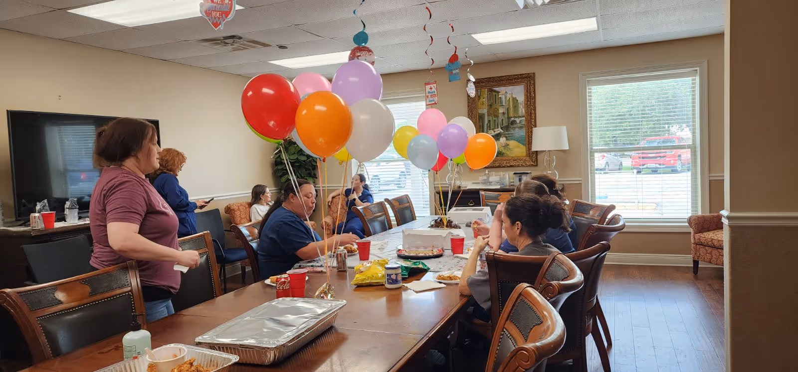 A group of people gathered around a long wooden table in a well-lit room decorated with colorful balloons. The table has food trays, snacks, and red cups. The room has large windows with blinds, a painting on the wall, and comfortable chairs. Some people are seated while others are standing and interacting.