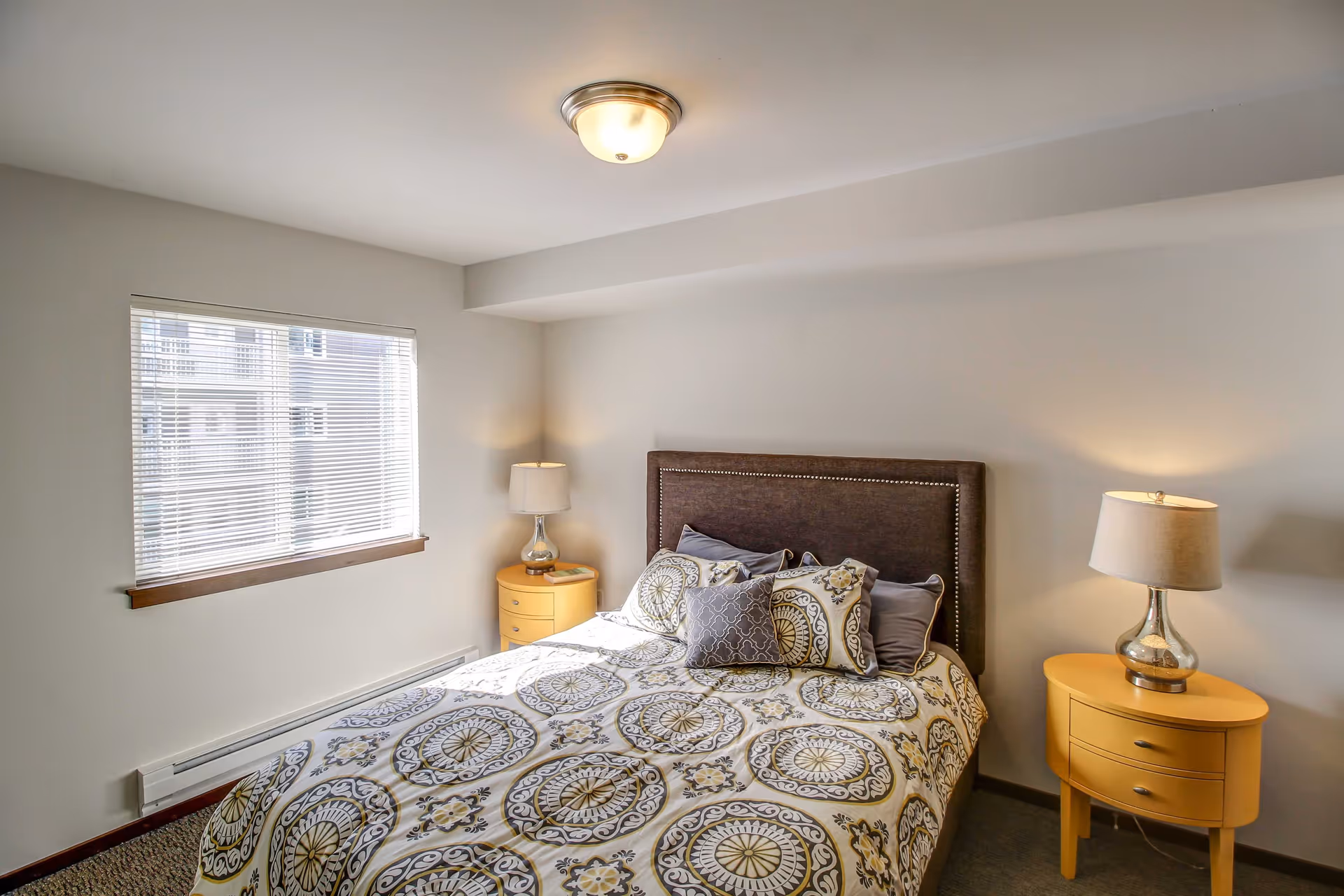 A bedroom with a bed featuring a patterned bedspread and multiple pillows. There are two yellow nightstands on either side of the bed, each with a lamp. A window with blinds is on the left wall, and the room has neutral-colored walls and ceiling with a ceiling light fixture.