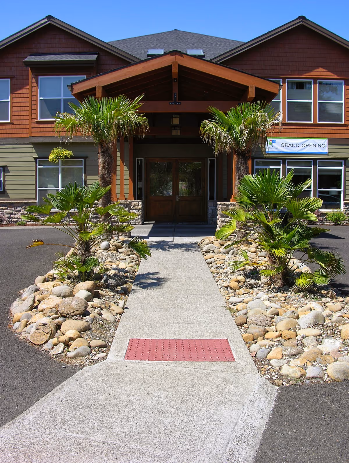Paved walkway leading to the front entrance of a two-story building flanked by palm plants and rock landscaping, with a 'Grand Opening' banner visible.