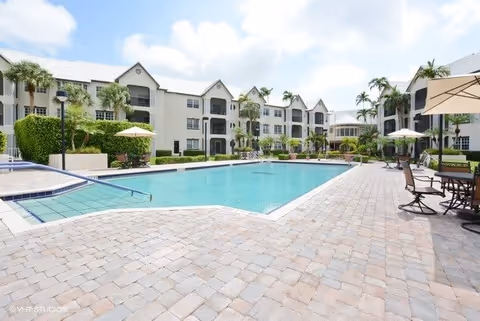 Outdoor swimming pool area surrounded by a paved deck with tables, chairs, and umbrellas. In the background, there is a multi-story residential building with balconies and palm trees under a partly cloudy sky.