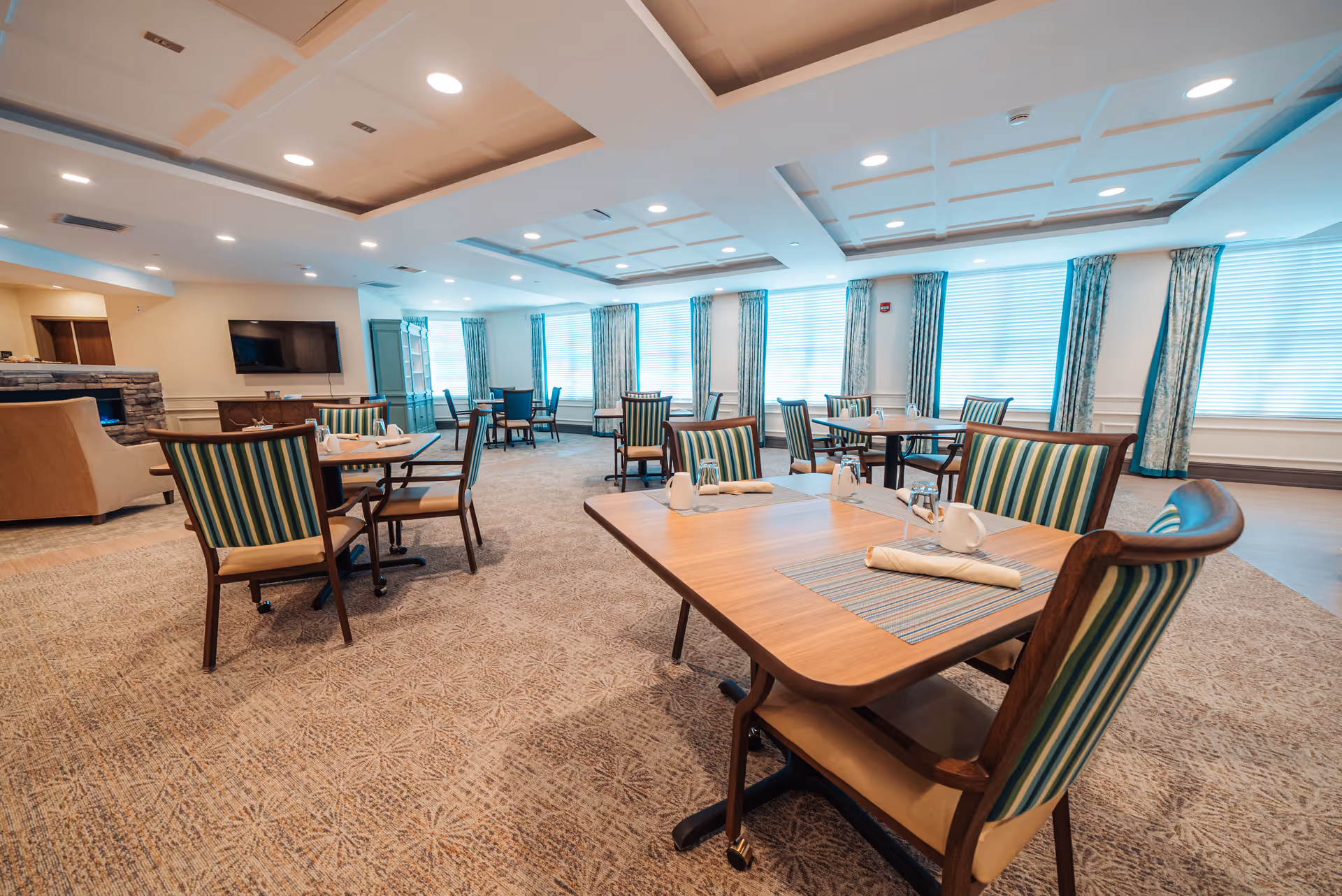 A spacious dining room with multiple wooden tables and chairs featuring striped upholstery. Each table is set with placemats, napkins, glasses, and salt and pepper shakers. The room has large windows with curtains allowing natural light to fill the space. There is a TV mounted on the wall and a fireplace with a stone surround in the background.