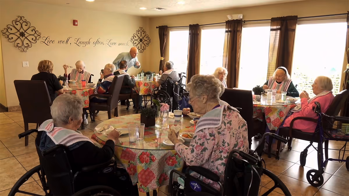 A group of elderly residents, some in wheelchairs, sitting around tables with floral tablecloths in a bright dining room. A staff member is serving or interacting with the residents. Large windows with brown curtains let in natural light. Wall decor includes a quote that reads 'Live well, Laugh often, Love much.'
