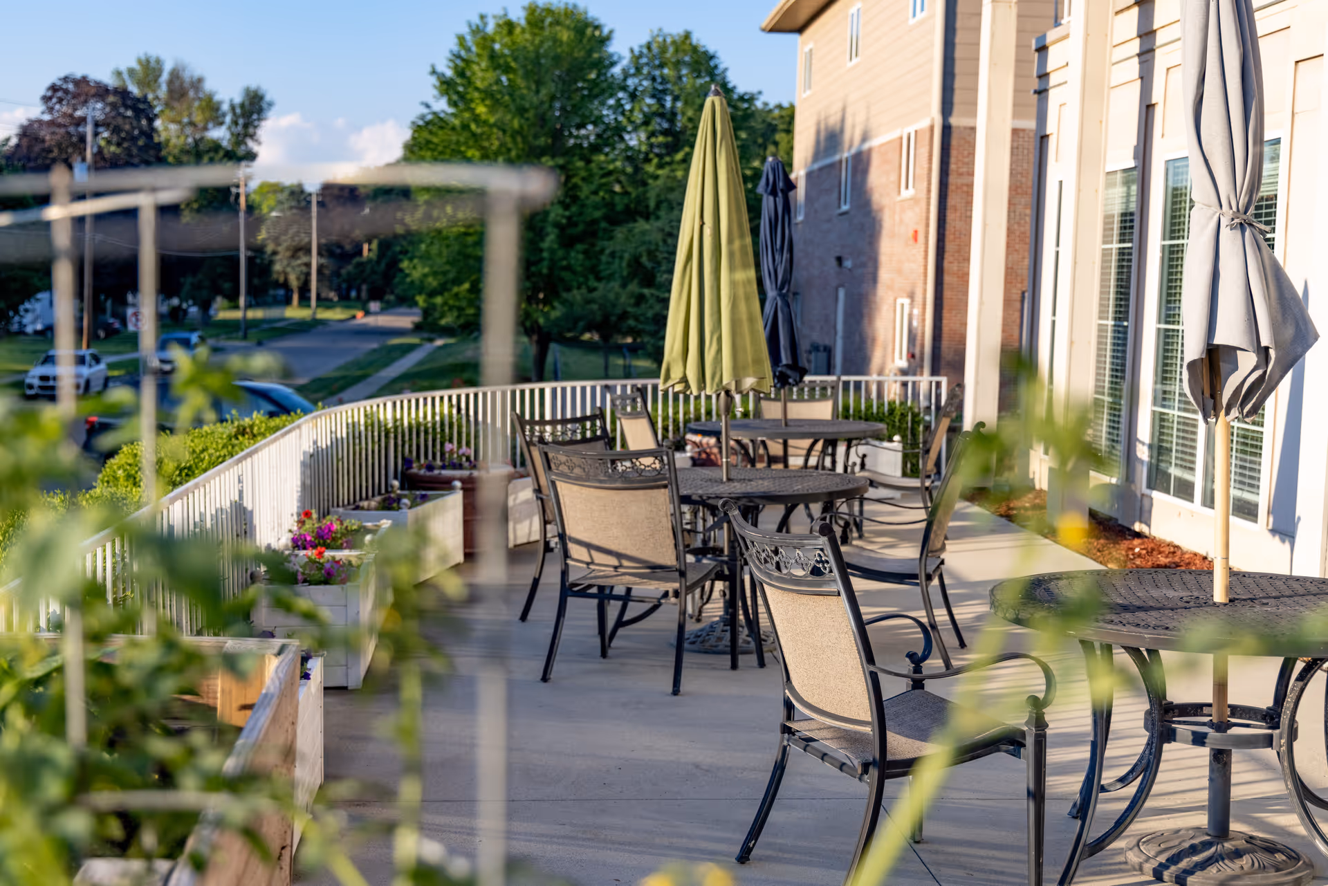 Outdoor patio area with several metal tables and chairs, some tables have closed umbrellas. The patio is adjacent to a building with large windows, and there are plants and flowers in planters along the railing. Trees and a street with parked cars are visible in the background under a clear sky.