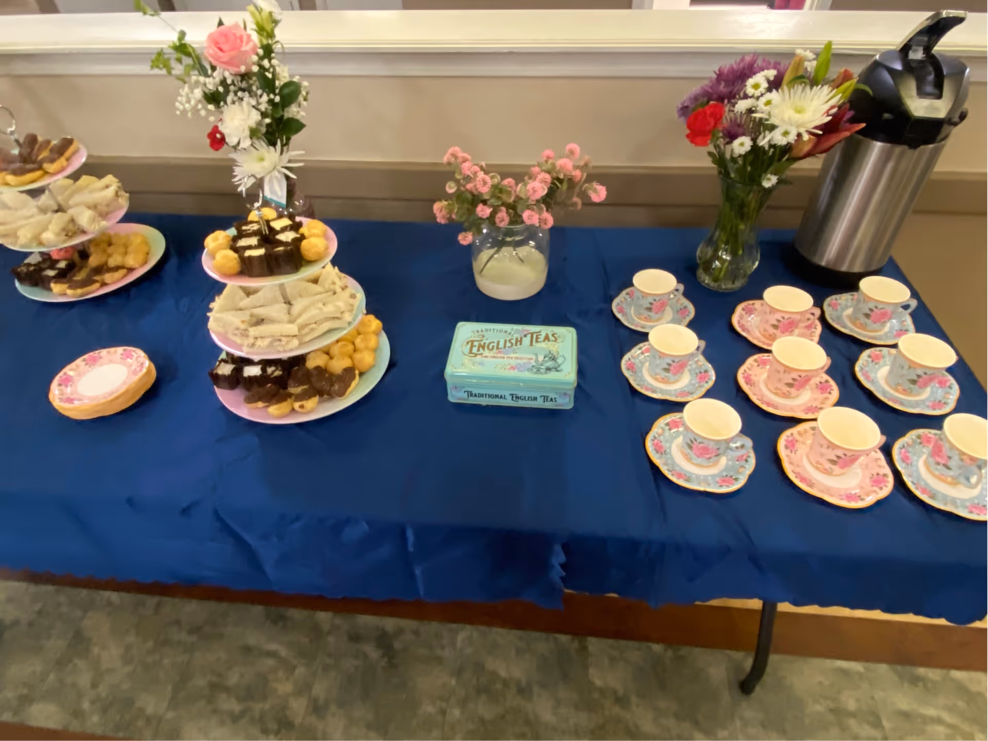 A table covered with a blue tablecloth displaying an assortment of tea party items including tiered trays with sandwiches, pastries, and desserts, floral arrangements in vases, a tin labeled 'Traditional English Teas,' several floral-patterned teacups and saucers, and a stainless steel airpot for hot beverages.