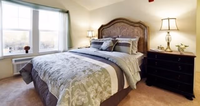 A cozy bedroom with a large bed featuring a decorative headboard, multiple pillows, and a patterned comforter. There are two bedside tables with lamps on each side of the bed. A window with curtains allows natural light to brighten the room.
