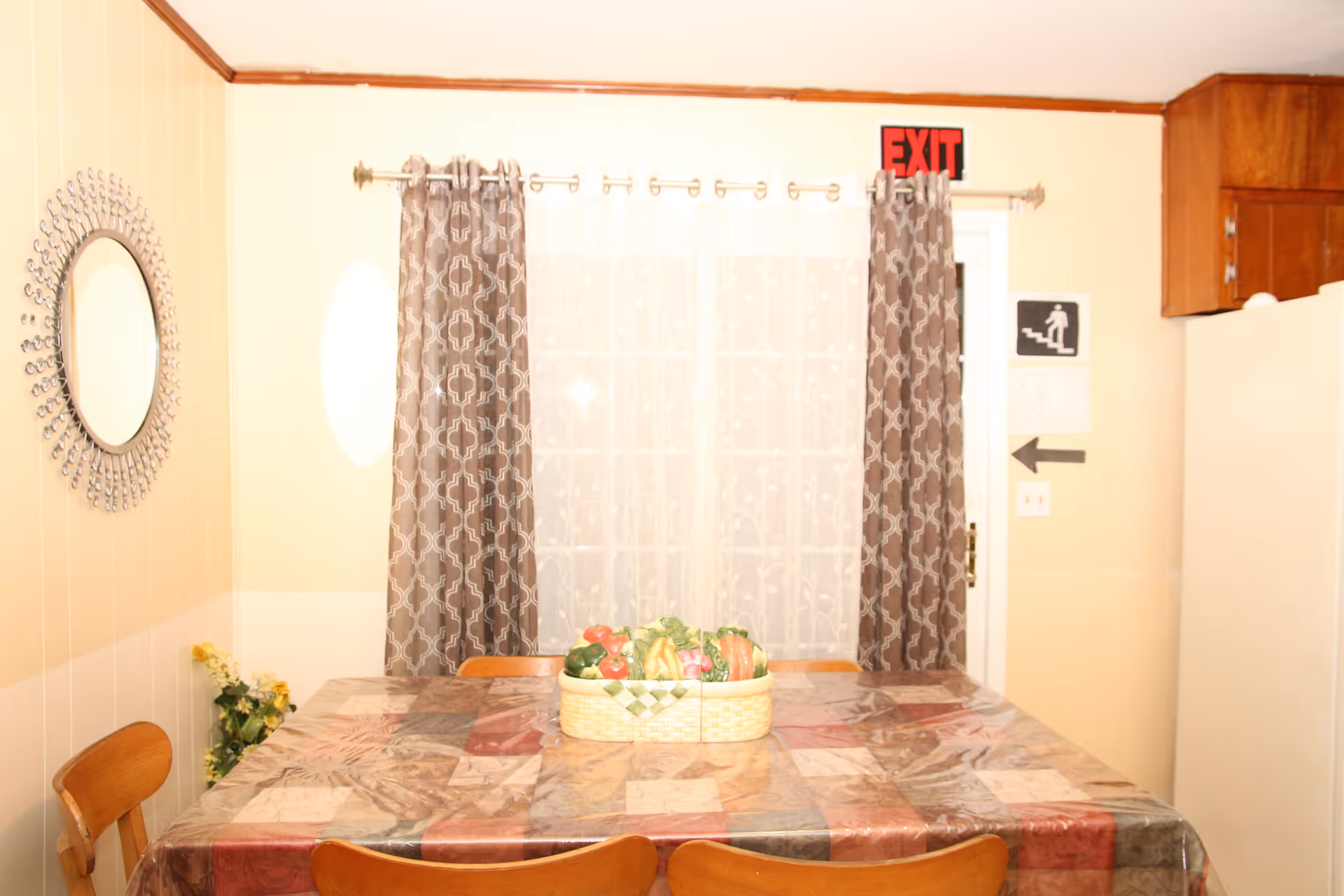 A dining area with a table covered by a patterned tablecloth and a plastic cover. On the table is a basket filled with artificial fruits and vegetables. Four wooden chairs surround the table. Behind the table is a window with sheer white curtains and patterned brown drapes. To the right, there is a white refrigerator and wooden cabinets above it. An exit sign is visible above a door with a stairway sign and an arrow pointing left. A decorative round mirror hangs on the left wall, and a small plant with yellow flowers is placed in the corner.