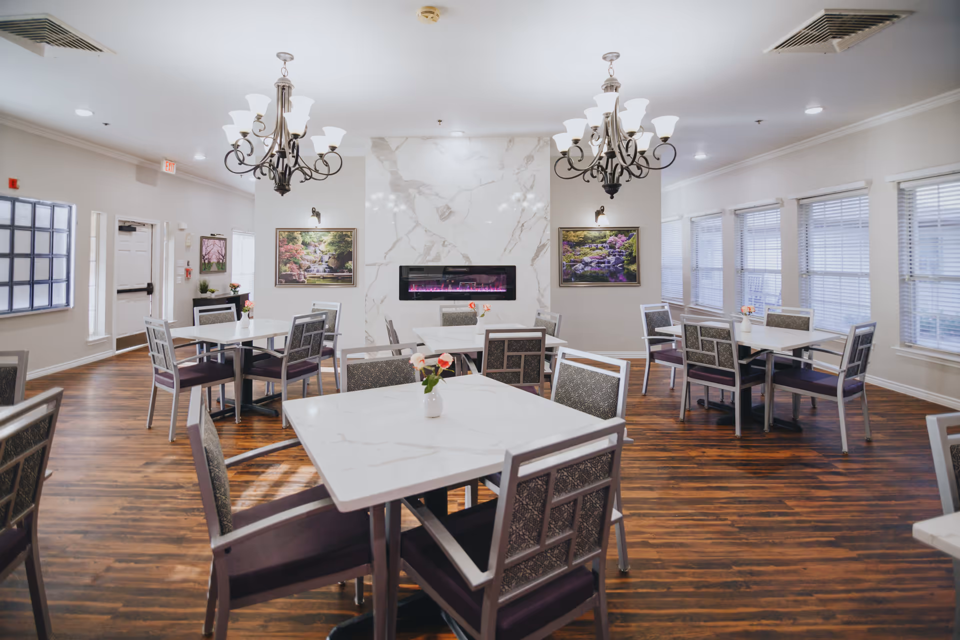 A bright and spacious dining room with several square white tables and cushioned chairs arranged neatly. The room features wooden flooring, two ornate chandeliers, a modern electric fireplace set in a marble wall, and framed nature paintings. Large windows with blinds allow natural light to fill the space.