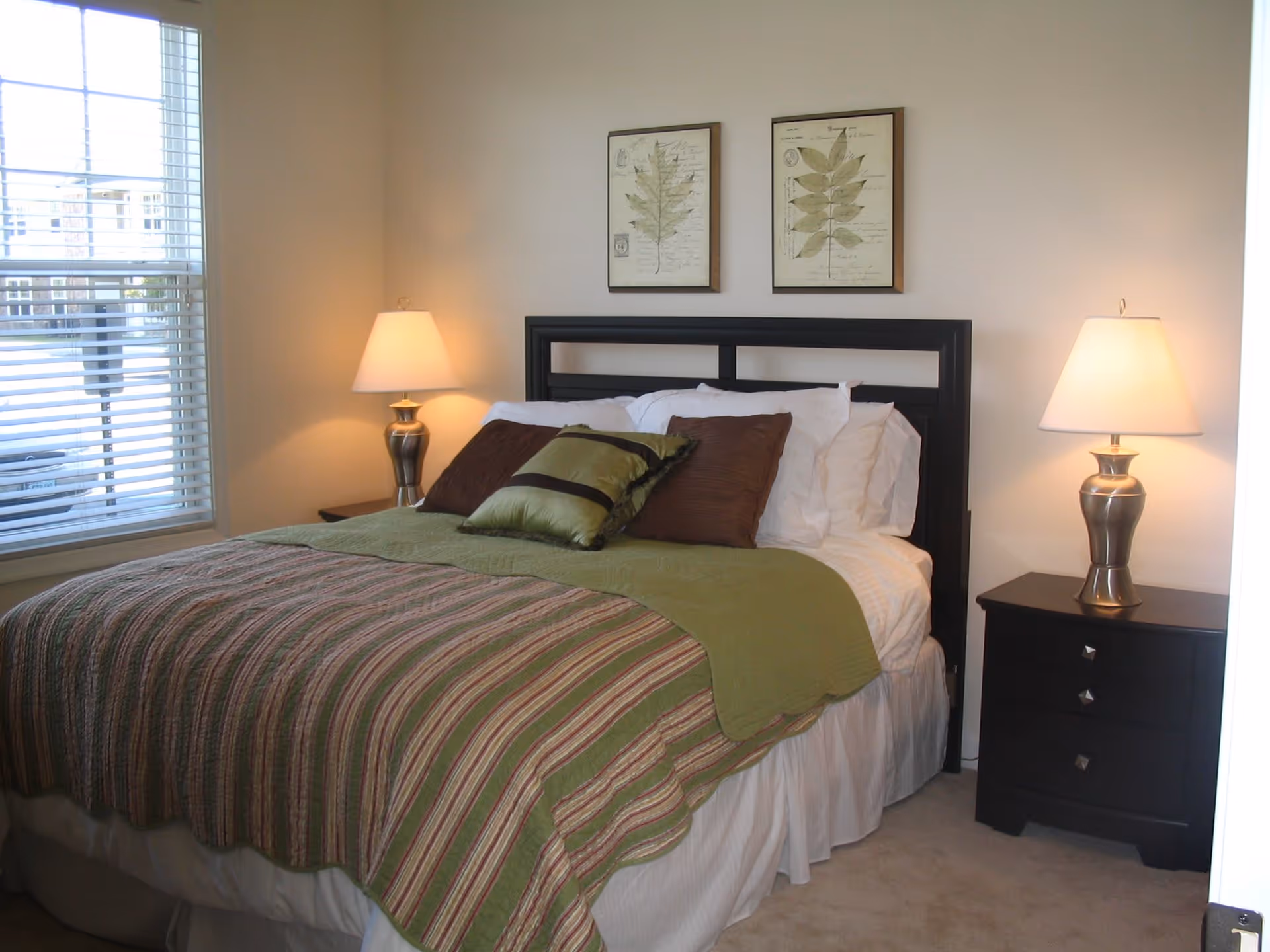A neatly made bed with a black headboard, green and brown striped bedspread, and multiple pillows in white, brown, and green. There are two bedside tables, each with a silver lamp with a white lampshade. Two framed botanical prints hang on the wall above the bed. A window with white blinds is visible on the left side of the room.