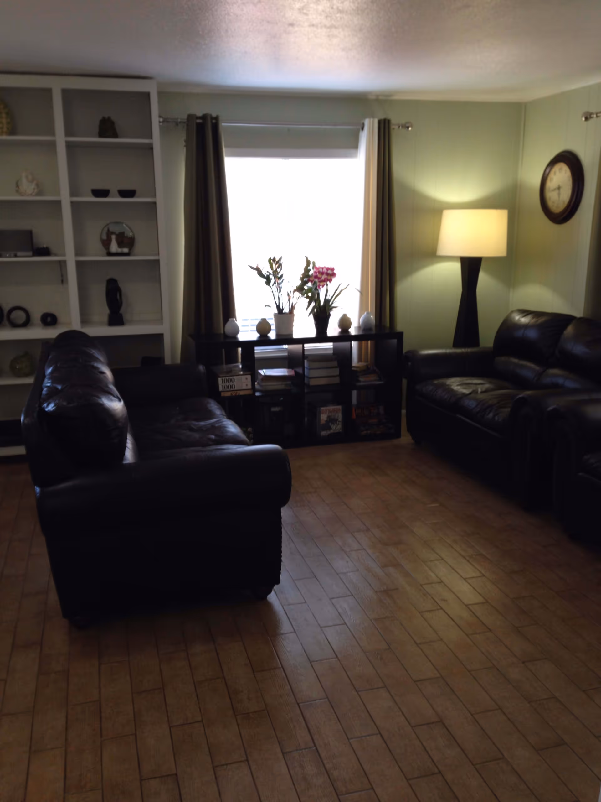 A cozy living room with two dark leather sofas facing each other, a wooden floor, a window with curtains, a black shelf with books and plants, a floor lamp, and a wall clock.