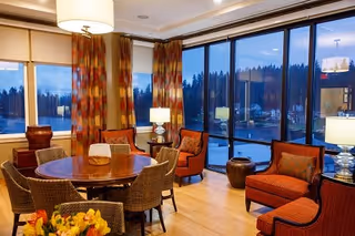 A cozy living room area with a round wooden table surrounded by wicker chairs and several cushioned armchairs. Large floor-to-ceiling windows provide a view of trees and the outdoors, with warm lighting from table lamps and a ceiling lamp creating a welcoming atmosphere.