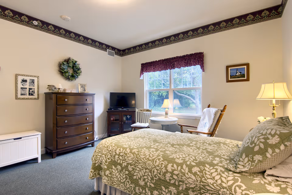 Bright furnished bedroom with a patterned green bedspread, wooden dresser and TV, a window with a valance, and a rocking chair beside a small table and lamp.