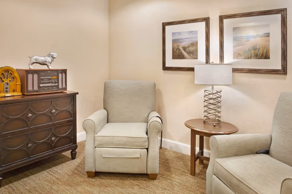 A cozy sitting area with two beige upholstered armchairs, a wooden side table with a lamp, and a dark wooden cabinet with a vintage radio and a silver dog figurine on top. Two framed landscape paintings hang on the beige wall behind the chairs.