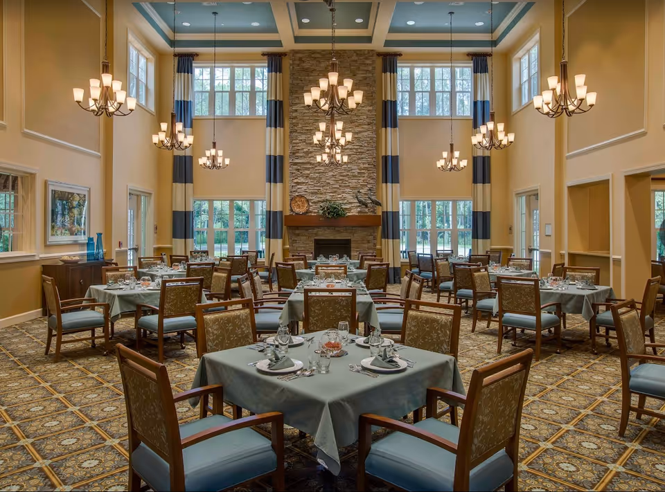 A spacious dining room with multiple tables covered in light blue tablecloths, each set with plates, glasses, and silverware. The room features high ceilings with several elegant chandeliers, large windows with blue and beige striped curtains, and a stone fireplace at the far end. The floor is carpeted with a patterned design, and the walls are painted in warm beige tones.